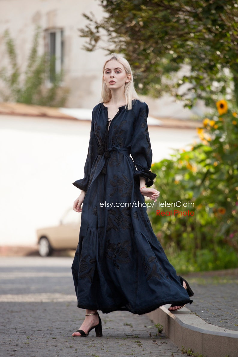 May include: A woman wearing a dark blue maxi dress with black embroidery. The dress has a V-neckline and long sleeves. The woman is standing on a sidewalk and looking at the camera. The text "etsy.com/shop/HelenCloth Real Photo" is visible in the image.