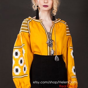 May include: A yellow embroidered blouse with black and white geometric patterns on the sleeves. The blouse has a V-neck and a long, flowing sleeve. The model is wearing a black skirt and has blonde hair. The text "etsy.com/shop/HelenCloth Real Photo" is visible at the bottom of the image.