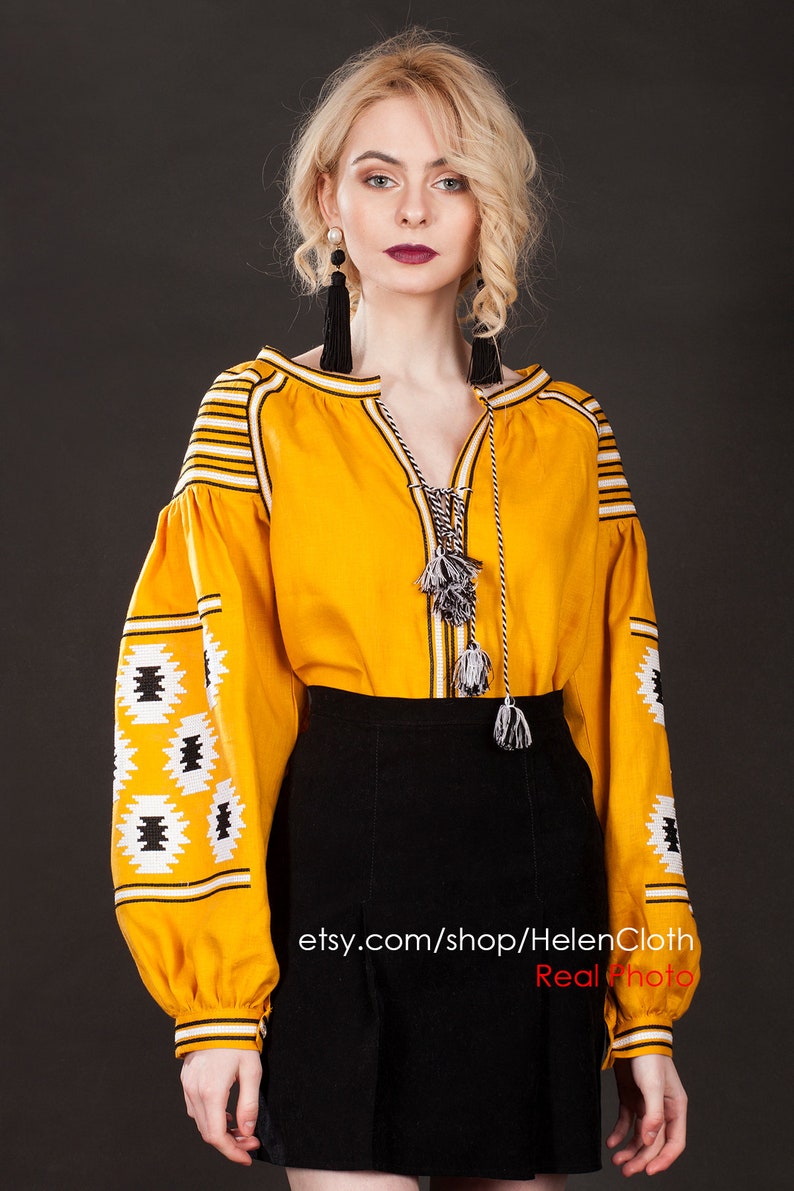 May include: A yellow embroidered blouse with black and white geometric patterns on the sleeves. The blouse has a V-neck and a long, flowing sleeve. The model is wearing a black skirt and has blonde hair. The text "etsy.com/shop/HelenCloth Real Photo" is visible at the bottom of the image.