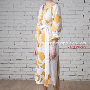 May include: A white linen dress with yellow embroidered floral patterns and tassels. The dress has a white sash and is worn by a person standing in front of a white brick wall.