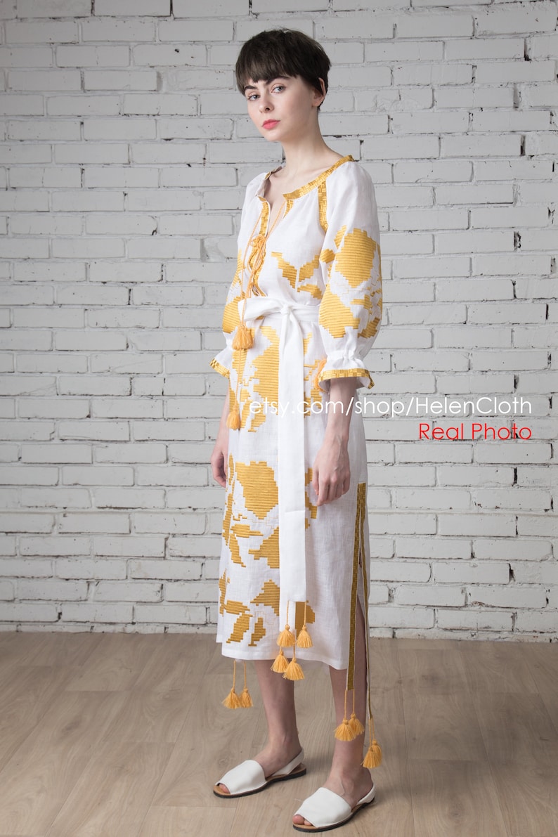 May include: A white linen dress with yellow embroidered floral patterns and tassels. The dress has a white sash and is worn by a person standing in front of a white brick wall.