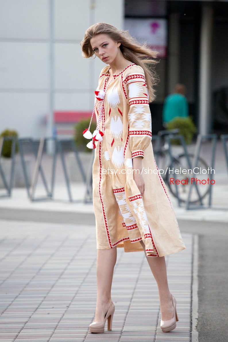 May include: A woman wearing a beige linen dress with red and white embroidery. The dress has a long sleeve and a loose fit. The woman is standing on a sidewalk with a brick pattern.