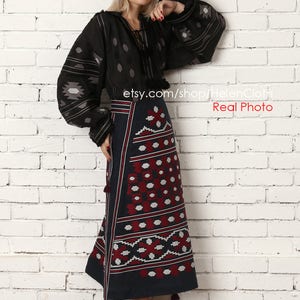 May include: A woman wearing a black embroidered blouse with long sleeves and a dark blue skirt with a red and white geometric pattern. The skirt has tassels on the bottom. She is standing in front of a white brick wall. The text "etsy.com/shop/HelenCloth Real Photo" is visible in the image.