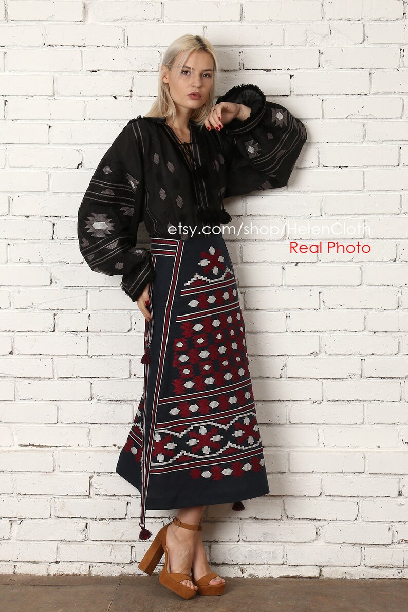 May include: A woman wearing a black embroidered blouse with long sleeves and a dark blue skirt with a red and white geometric pattern. The skirt has tassels on the bottom. She is standing in front of a white brick wall. The text "etsy.com/shop/HelenCloth Real Photo" is visible in the image.