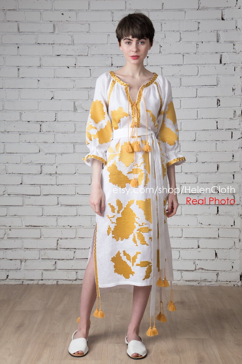 May include: A white linen dress with yellow embroidered floral patterns. The dress has a V-neckline, 3/4 sleeves, and a tie at the waist. The dress is shown on a model with a white brick wall background.
