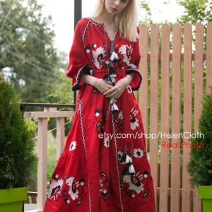 May include: A woman wearing a red embroidered linen dress with black and white floral patterns. The dress has a ruffled hem and a belt. The woman is standing in front of a wooden fence and has her hands clasped in front of her. The text "etsy.com/shop/HelenCloth Real Photo" is visible in the image.