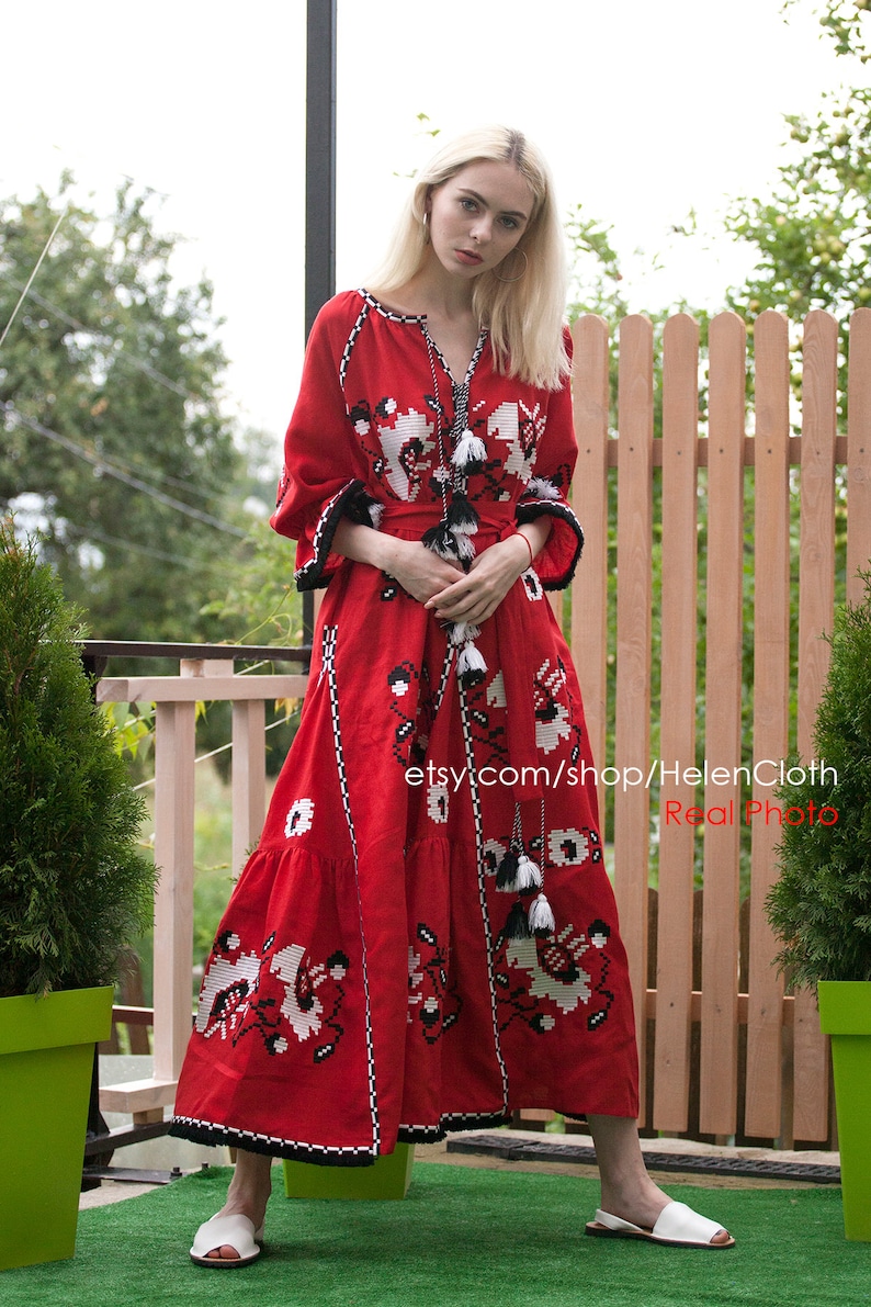May include: A woman wearing a red embroidered linen dress with black and white floral patterns. The dress has a ruffled hem and a belt. The woman is standing in front of a wooden fence and has her hands clasped in front of her. The text "etsy.com/shop/HelenCloth Real Photo" is visible in the image.