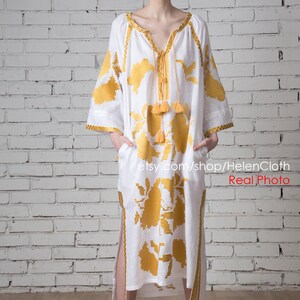 May include: A white linen dress with yellow embroidered floral patterns and tassels. The dress has a V-neckline and long sleeves. The model is wearing white sandals. Real Photo etsy.com/shop/HelenCloth
