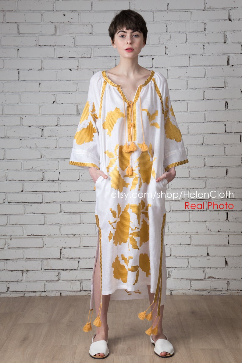 May include: A white linen dress with yellow embroidered floral patterns and tassels. The dress has a V-neckline and long sleeves. The model is wearing white sandals. Real Photo etsy.com/shop/HelenCloth