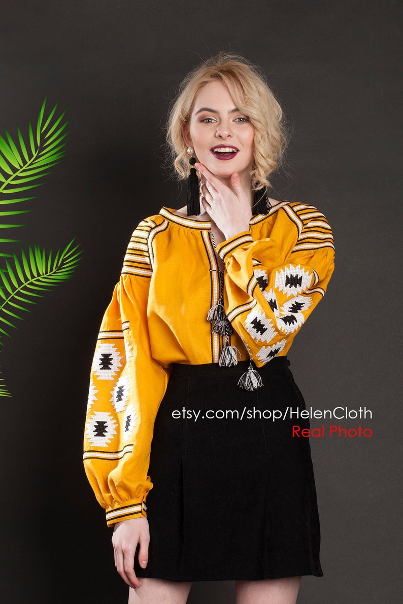 May include: A yellow embroidered blouse with black and white geometric patterns. The blouse has long sleeves with a gathered cuff and a round neckline. The model is wearing a black skirt and has blonde hair.