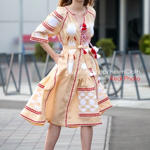 May include: A woman wearing a beige linen dress with white and red embroidery. The dress has a cinched waist and a flowing skirt. The dress is decorated with tassels and has a traditional Ukrainian design.