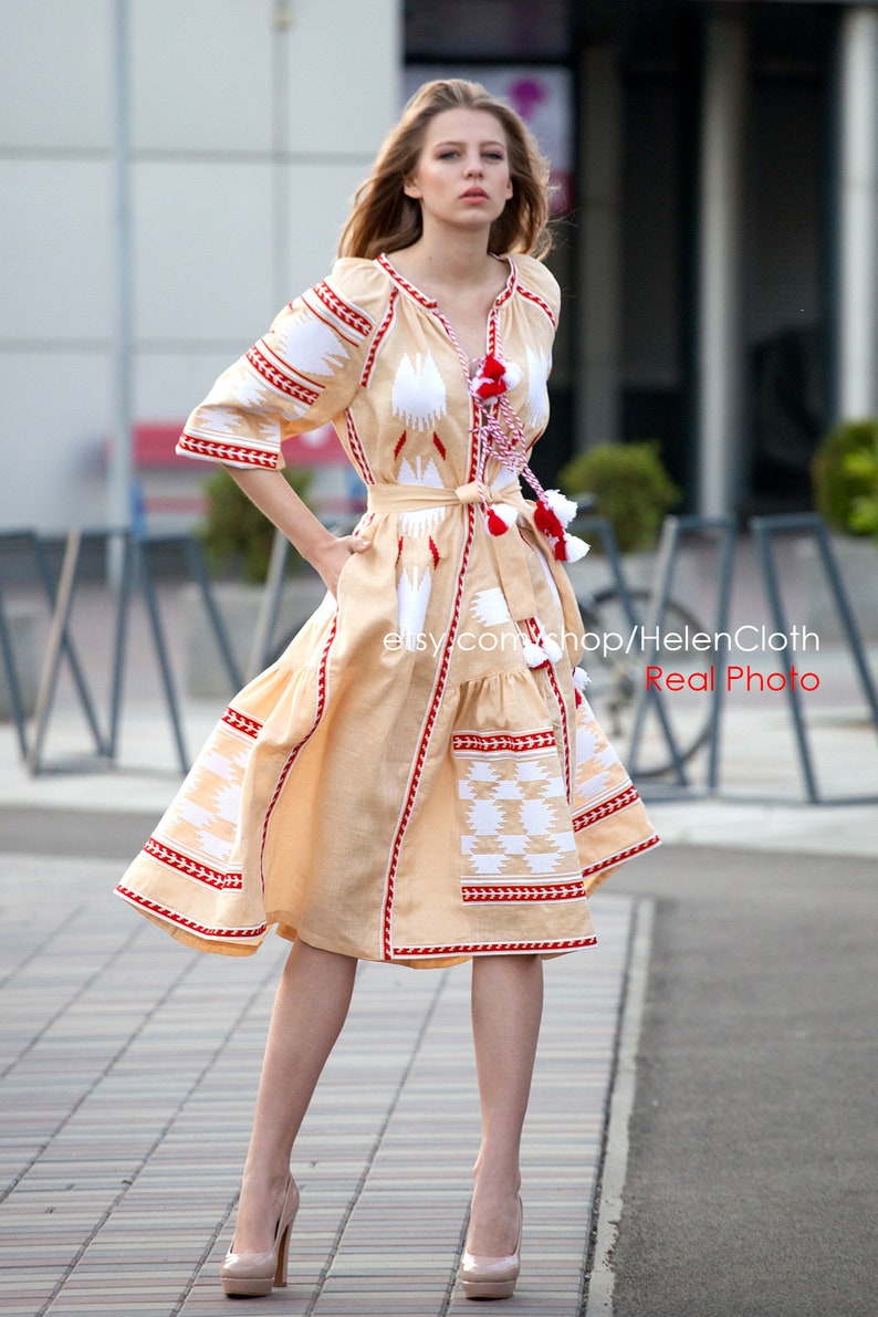 May include: A woman wearing a beige linen dress with white and red embroidery. The dress has a cinched waist and a flowing skirt. The dress is decorated with tassels and has a traditional Ukrainian design.