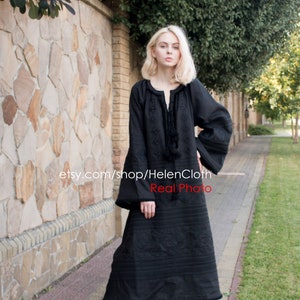 May include: A black embroidered linen dress with long sleeves and a round neckline. The dress has a loose fit and falls to the ankles. The model is wearing white sneakers.