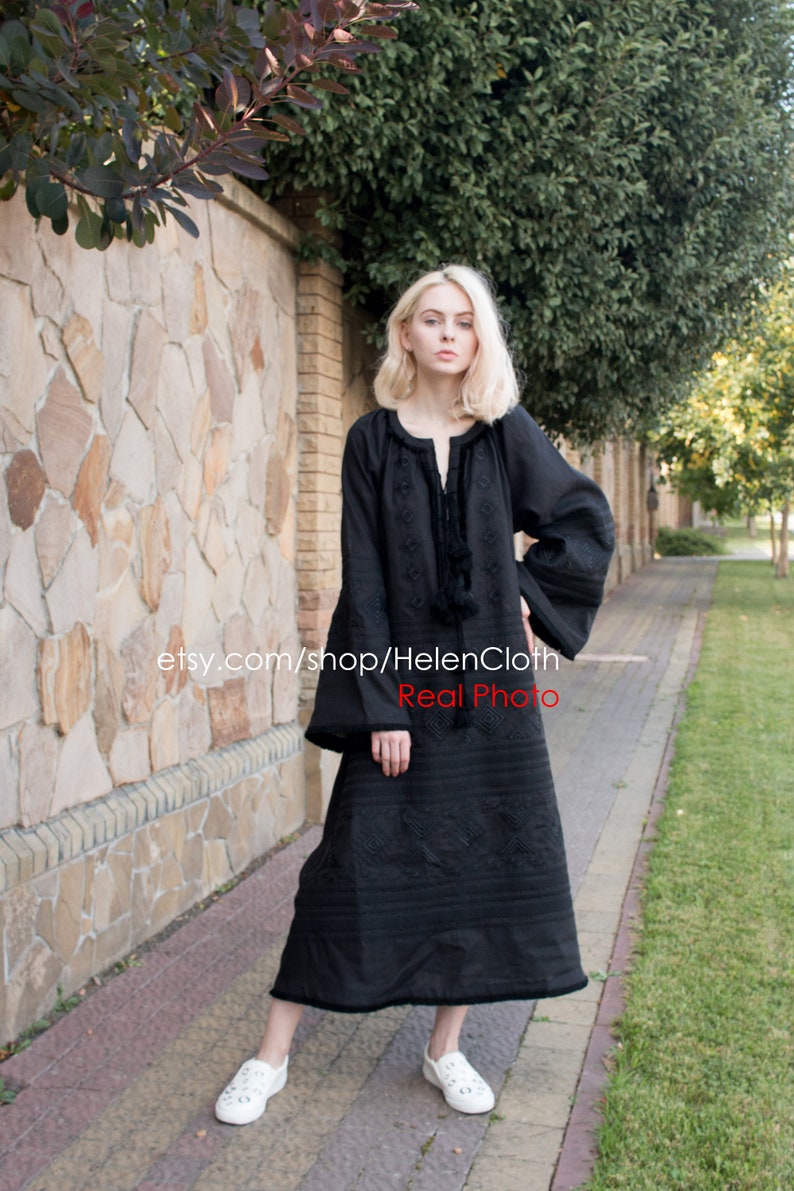 May include: A black embroidered linen dress with long sleeves and a round neckline. The dress has a loose fit and falls to the ankles. The model is wearing white sneakers.