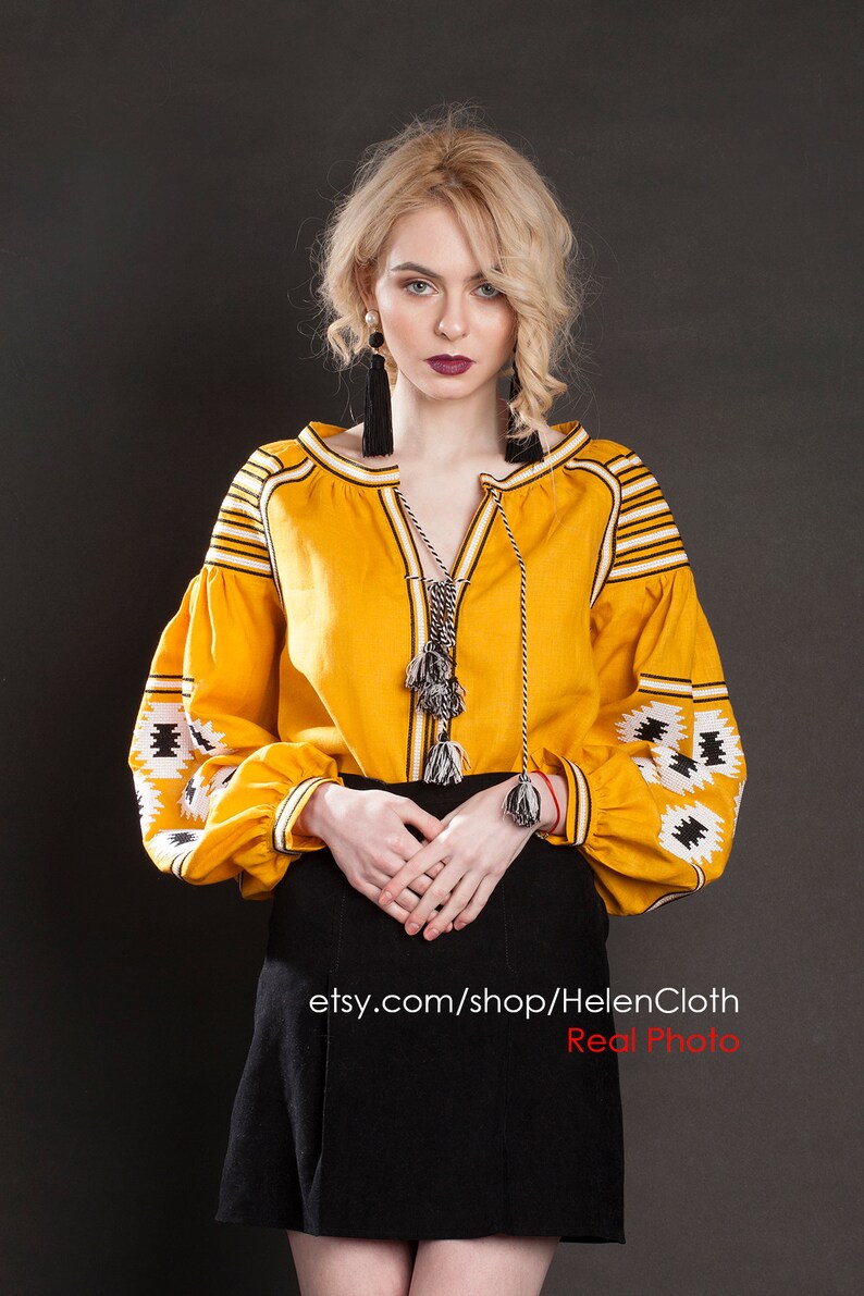 May include: A yellow embroidered linen blouse with black and white geometric patterns. The blouse has a V-neckline and long sleeves with a tassel detail. The model is wearing a black skirt and has blonde hair. The text "etsy.com/shop/HelenCloth Real Photo" is visible at the bottom of the image.