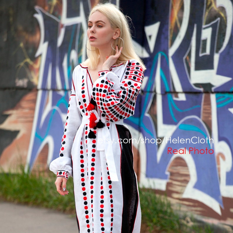 May include: A white linen dress with red and black polka dots. The dress has a long sleeve and a V-neck. The dress is belted at the waist and has a black and white trim. The dress is worn by a woman with blonde hair and a white background.