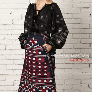 May include: A black and white embroidered blouse with long sleeves and a navy blue skirt with red and white geometric patterns. The skirt has pockets and tassels on the bottom.