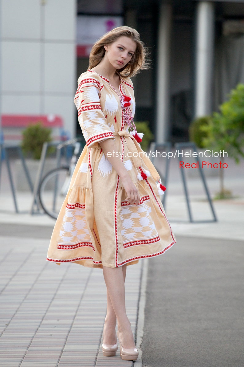 May include: A beige embroidered dress with red and white trim. The dress has a fitted bodice and a flowing skirt. The model is wearing the dress with a belt and has her hair pulled back.