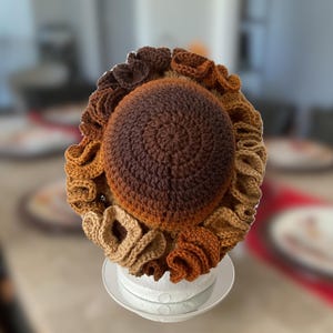 May include: A crocheted sunflower cushion with brown, tan and orange petals. The centre of the sunflower is a brown and tan ombre colour.