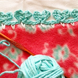 May include: A close-up of a teal crochet edging being added to a pink and white fleece blanket. A ball of teal yarn and a crochet hook are also visible.