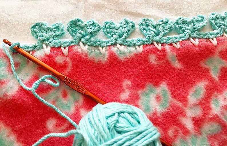 May include: A close-up of a teal crochet edging being added to a pink and white fleece blanket. A ball of teal yarn and a crochet hook are also visible.