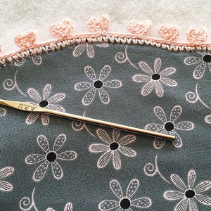 May include: A close-up of a crochet hook with a pink crocheted edging on a fabric with a floral pattern. The crochet hook is labeled "4.25"