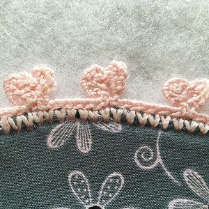 May include: Close-up of a pink crocheted trim with three flower-shaped embellishments. The trim is attached to a gray fabric with a floral pattern.