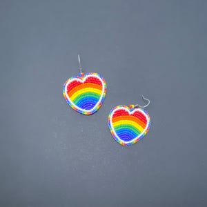 May include: Two heart-shaped earrings with a rainbow design. The earrings are made of small, colorful beads arranged in a heart shape, with a white border and silver hooks. The rainbow colors are red, orange, yellow, green, blue, and purple.