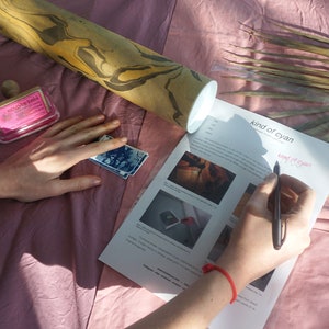 Puede incluir: Una persona est&aacute; escribiendo en una hoja de papel con el texto "kind of cyan" impreso en ella. El papel est&aacute; sobre una superficie rosa con un rodillo de madera, un sello rosa y una hoja de palma seca.