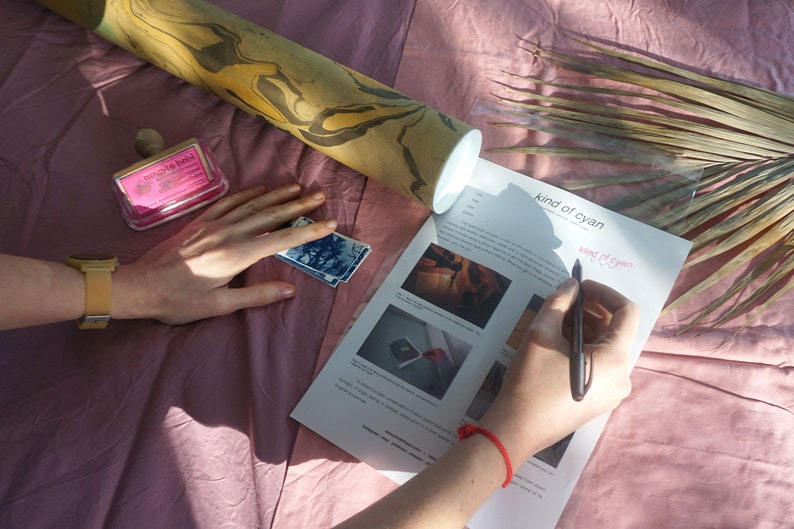 Puede incluir: Una persona est&aacute; escribiendo en una hoja de papel con el texto "kind of cyan" impreso en ella. El papel est&aacute; sobre una superficie rosa con un rodillo de madera, un sello rosa y una hoja de palma seca.