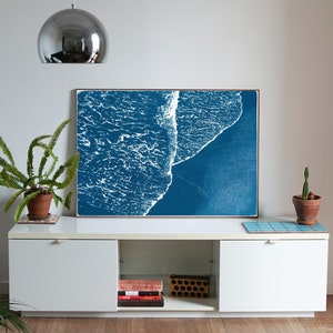 Puede incluir: Una impresi&oacute;n enmarcada de un dise&ntilde;o abstracto de ola de oc&eacute;ano azul y blanco. La impresi&oacute;n est&aacute; colgada en una pared blanca sobre un armario blanco con una planta de helecho a la izquierda y una planta de cactus a la derecha.