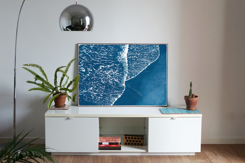 Puede incluir: Una impresi&oacute;n enmarcada de un dise&ntilde;o abstracto de ola de oc&eacute;ano azul y blanco. La impresi&oacute;n est&aacute; colgada en una pared blanca sobre un armario blanco con una planta de helecho a la izquierda y una planta de cactus a la derecha.