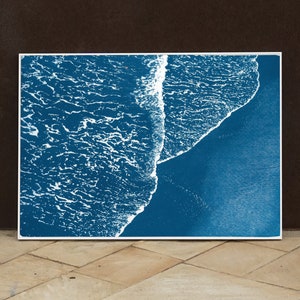 Puede incluir: Una fotograf&iacute;a a&eacute;rea abstracta de una ola rompiendo en una playa. La imagen est&aacute; en tonos azules y blancos, con la ola apareciendo como una l&iacute;nea blanca contra el agua azul.