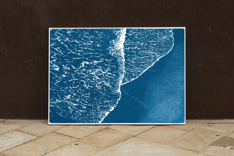 Puede incluir: Una fotograf&iacute;a a&eacute;rea abstracta de una ola rompiendo en una playa. La imagen est&aacute; en tonos azules y blancos, con la ola apareciendo como una l&iacute;nea blanca contra el agua azul.