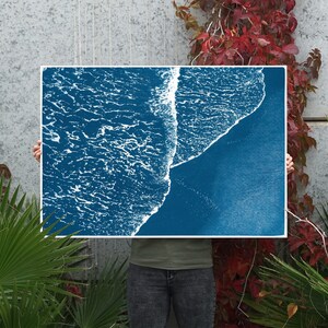 Puede incluir: Una impresi&oacute;n azul cian de una ola rompiendo en una playa. La impresi&oacute;n la sostiene una persona frente a una pared gris con plantas verdes.
