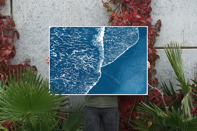 Puede incluir: Una impresi&oacute;n azul cian de una ola rompiendo en una playa. La impresi&oacute;n la sostiene una persona frente a una pared gris con plantas verdes.