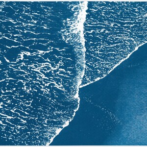 Puede incluir: Vista a&eacute;rea de una ola rompiendo en una playa, creando una l&iacute;nea blanca espumosa contra el agua azul oscuro.