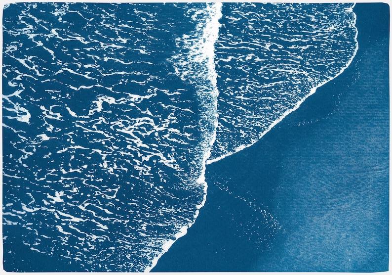 Puede incluir: Vista a&eacute;rea de una ola rompiendo en una playa, creando una l&iacute;nea blanca espumosa contra el agua azul oscuro.
