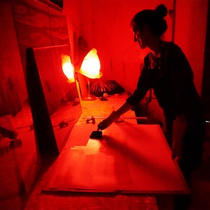 Puede incluir: Una persona en una habitaci&oacute;n oscura utiliza un pincel para aplicar un producto qu&iacute;mico a un papel fotogr&aacute;fico bajo una luz roja de seguridad.