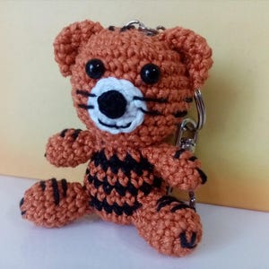 Peut inclure: Un porte-clés tigre crocheté orange et noir. Le tigre a des rayures noires, des yeux noirs et un museau blanc et noir. Le porte-clés est attaché à un anneau en métal argenté.