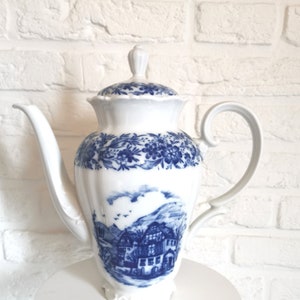 Puede incluir: Una tetera de porcelana blanca con un diseño floral azul y blanco y una escena de paisaje azul y blanco. La tetera tiene una tapa y un asa.