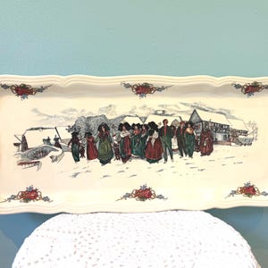 Könnte beinhalten: Cremefarbene rechteckige Servierplatte mit gewelltem Rand. Sie zeigt eine Winterszene mit Figuren in traditioneller Kleidung und Häusern. Rote und blaue Blumenmotive verzieren die Ecken.