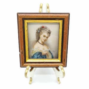 Può includere: Un ritratto in miniatura incorniciato di una donna con un vestito blu, una collana di perle e un fiore nei capelli. La cornice è in legno con una bordura dorata.