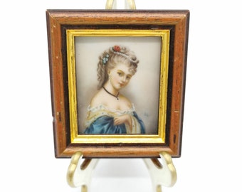 May include: A framed miniature portrait of a woman in a blue dress with a pearl necklace and a flower in her hair. The frame is made of wood with a gold trim.