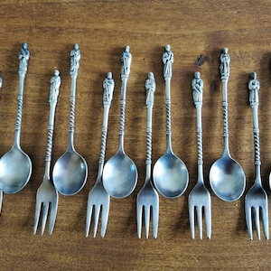 May include: A collection of antique silver-toned spoons and forks. Each spoon and fork has a decorative handle with a twisted design and a small statue at the top. The utensils are arranged on a wooden surface.