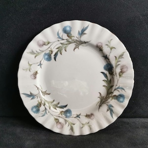 Puede incluir: Plato de cerámica blanca con borde ondulado, decorado con un motivo floral de cardos azules y morados y hojas verdes. El plato tiene un diseño clásico.