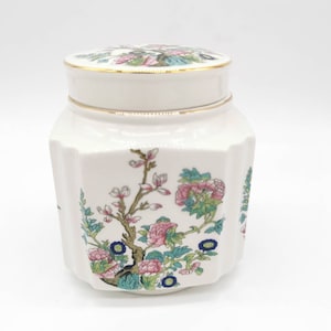 Può includere: Vaso in ceramica bianca con bordo dorato e motivo floreale. Il vaso ha un coperchio con un motivo floreale. Il vaso è ottagonale.