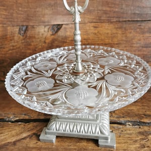 Pode incluir: Um suporte para bolo de prata e vidro com uma alça decorativa. O suporte tem uma base quadrada e uma parte superior redonda de vidro com um design de vidro cortado.