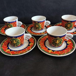 May include: Set of five espresso cups and saucers. The cups have white interiors and wing-shaped handles. The saucers and cups are decorated with a red, green, blue, and gold ornate design. The set is on a dark surface.