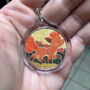May include: A circular clear acrylic keychain featuring a printed illustration of a stylized orange fox-like creature with multiple tails, standing amidst pink roses and a yellow background with small star accents, attached to a silver metal ring.